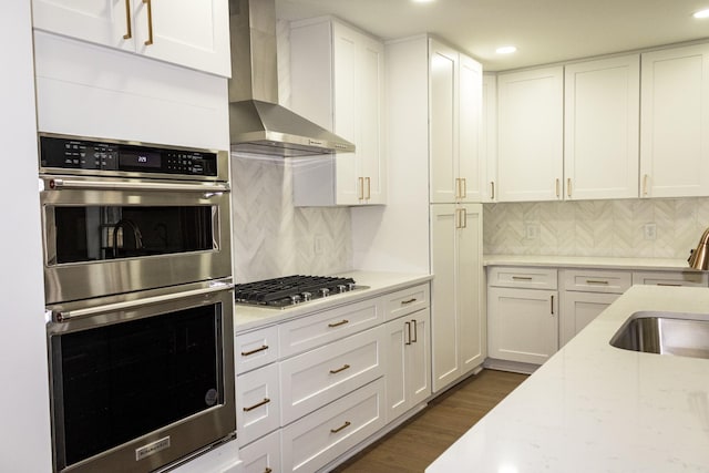 kitchen featuring wall chimney range hood, sink, stainless steel appliances, light stone counters, and white cabinets