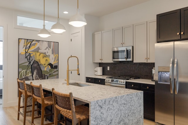 kitchen featuring an island with sink, stainless steel appliances, tasteful backsplash, decorative light fixtures, and sink