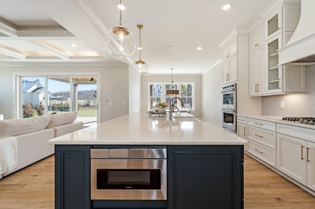 kitchen with white cabinetry, custom exhaust hood, hanging light fixtures, and an island with sink