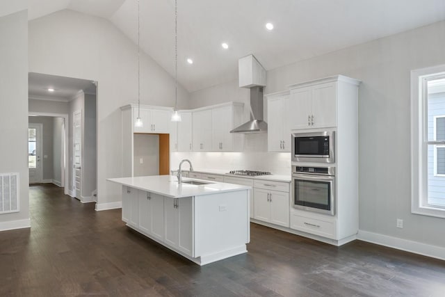 kitchen featuring appliances with stainless steel finishes, wall chimney exhaust hood, decorative light fixtures, white cabinetry, and a center island with sink