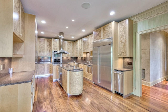 kitchen with an island with sink, island exhaust hood, stainless steel appliances, decorative backsplash, and light wood-type flooring