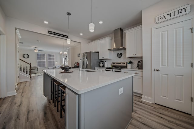 kitchen featuring pendant lighting, appliances with stainless steel finishes, a kitchen island with sink, white cabinetry, and wall chimney exhaust hood