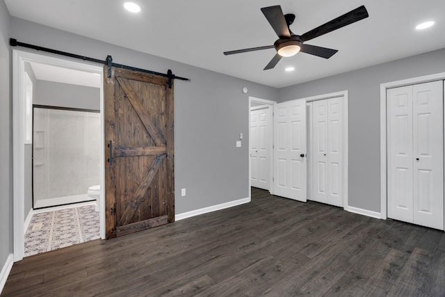 unfurnished bedroom featuring ceiling fan, a barn door, two closets, dark wood-type flooring, and connected bathroom