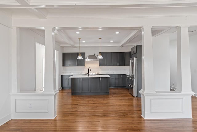 kitchen featuring hanging light fixtures, beam ceiling, an island with sink, and wall chimney range hood