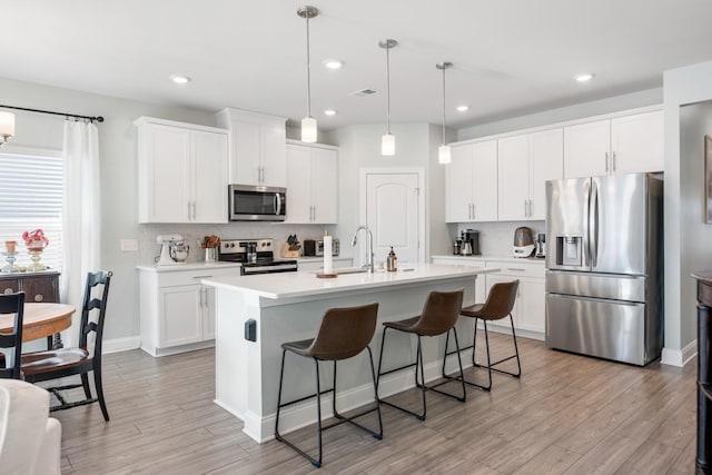 kitchen with appliances with stainless steel finishes, white cabinetry, tasteful backsplash, hanging light fixtures, and a kitchen island with sink