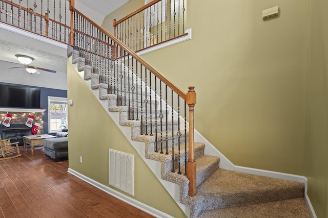 staircase featuring ceiling fan, a towering ceiling, a stone fireplace, and hardwood / wood-style floors