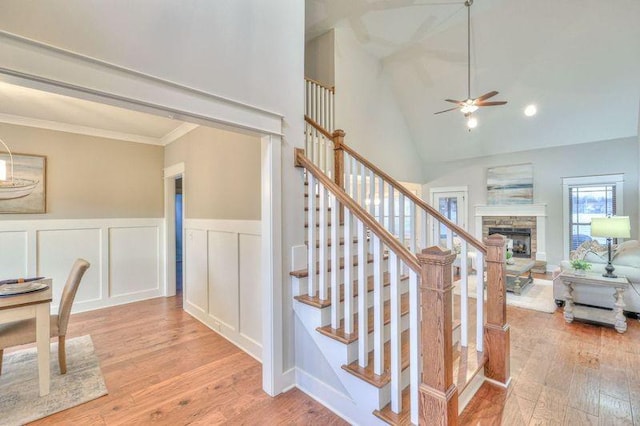 staircase featuring a towering ceiling, a stone fireplace, ceiling fan, ornamental molding, and hardwood / wood-style flooring