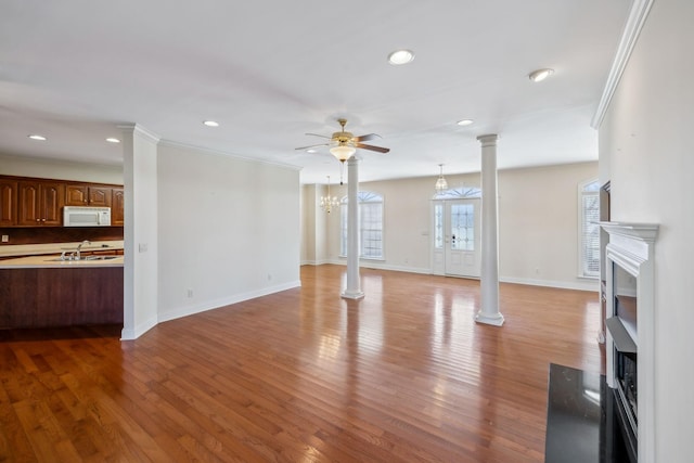 unfurnished living room featuring sink, crown molding, light wood-type flooring, ceiling fan with notable chandelier, and decorative columns