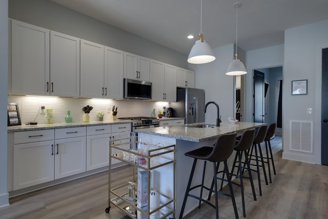 kitchen with a center island with sink, white cabinetry, hanging light fixtures, appliances with stainless steel finishes, and light stone counters