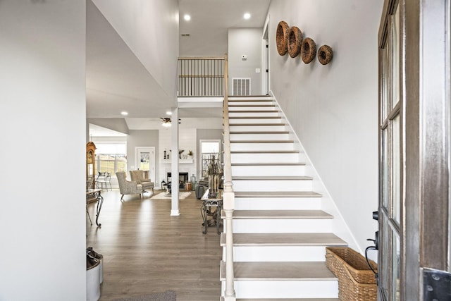 stairs with ceiling fan, a large fireplace, hardwood / wood-style flooring, and a towering ceiling