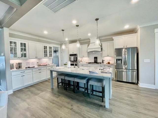 kitchen with stainless steel appliances, custom exhaust hood, white cabinets, and an island with sink