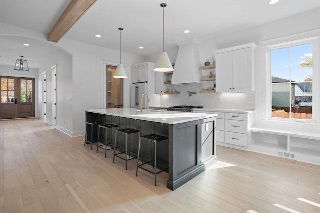 kitchen featuring decorative light fixtures, beamed ceiling, white cabinetry, a large island with sink, and custom range hood