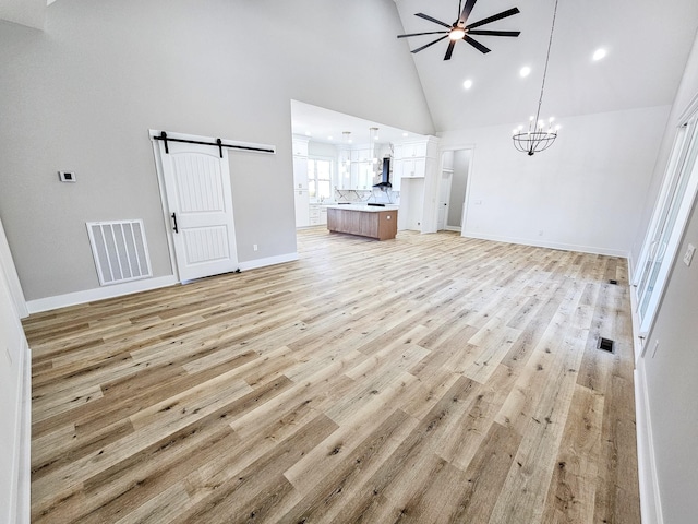 unfurnished living room with light wood-type flooring, ceiling fan with notable chandelier, a barn door, and high vaulted ceiling