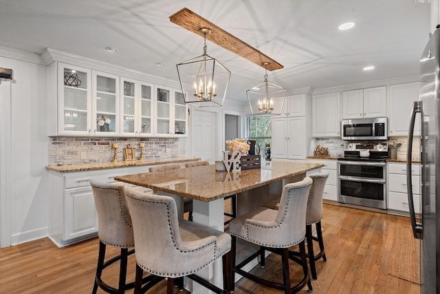 kitchen with white cabinetry, stainless steel appliances, hanging light fixtures, a kitchen island with sink, and light stone counters