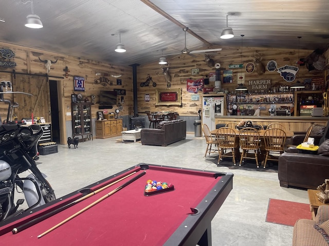 recreation room featuring ceiling fan, vaulted ceiling, concrete floors, wooden walls, and a wood stove