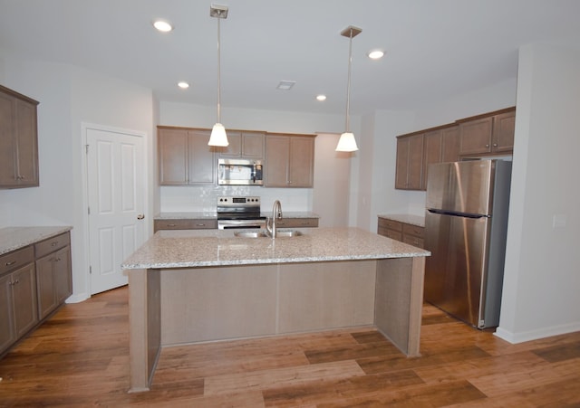 kitchen with a center island with sink, sink, hanging light fixtures, appliances with stainless steel finishes, and light stone counters