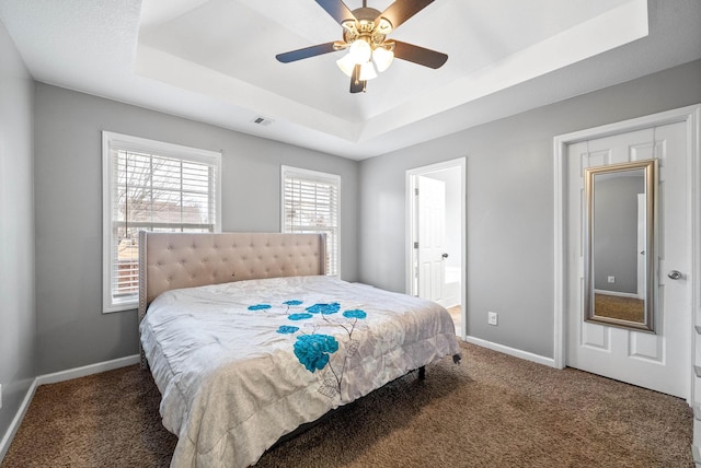 bedroom with ceiling fan, dark colored carpet, a tray ceiling, and ensuite bath