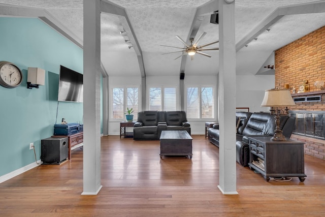 living room with ceiling fan, a fireplace, light hardwood / wood-style floors, lofted ceiling with beams, and a textured ceiling