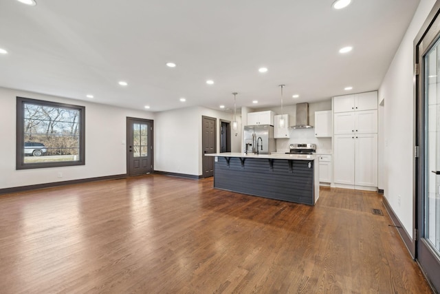 kitchen with a center island with sink, appliances with stainless steel finishes, pendant lighting, wall chimney exhaust hood, and white cabinets