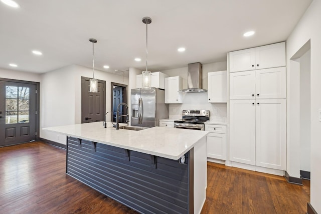 kitchen featuring appliances with stainless steel finishes, an island with sink, decorative light fixtures, and wall chimney range hood