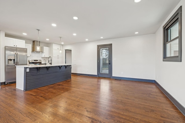 kitchen with decorative light fixtures, wall chimney range hood, a kitchen island with sink, stainless steel appliances, and white cabinets