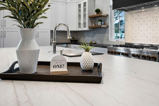 interior details with tasteful backsplash, light stone countertops, stainless steel gas cooktop, white cabinets, and wall chimney exhaust hood