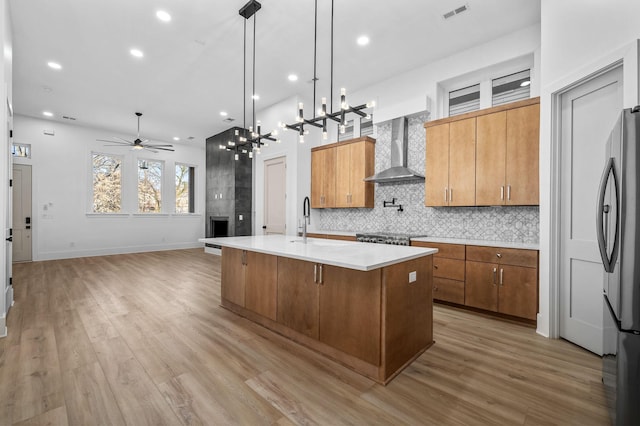 kitchen with light hardwood / wood-style floors, stainless steel refrigerator, hanging light fixtures, an island with sink, and wall chimney exhaust hood