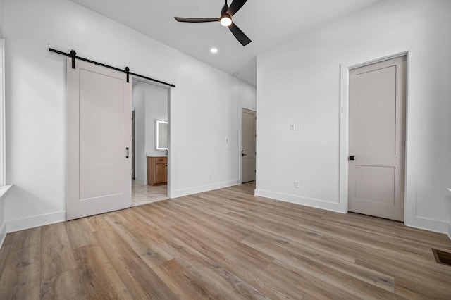 unfurnished bedroom featuring ceiling fan, ensuite bathroom, a barn door, and light hardwood / wood-style flooring