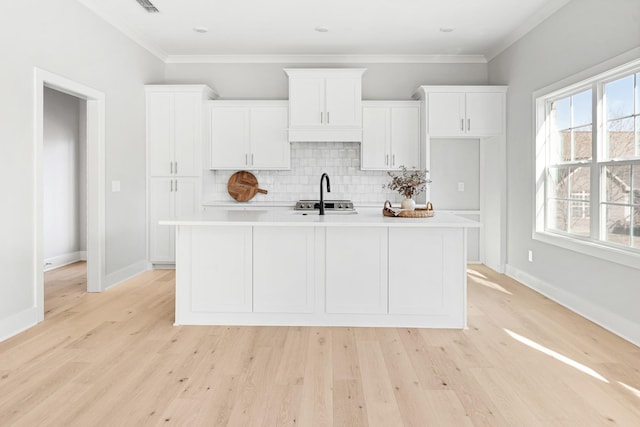 kitchen with white cabinets, backsplash, a kitchen island with sink, crown molding, and light wood-type flooring