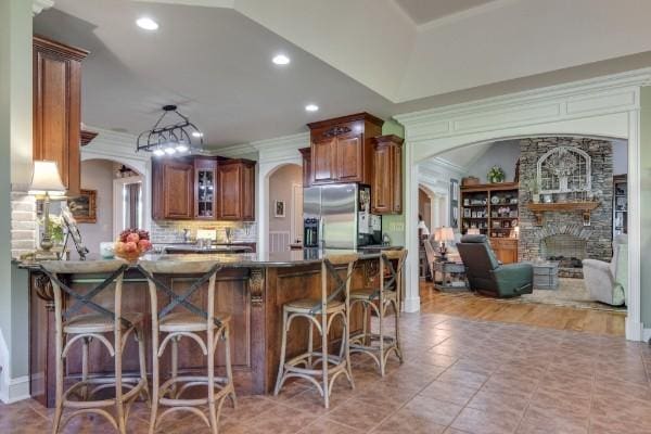 kitchen with a fireplace, kitchen peninsula, stainless steel refrigerator with ice dispenser, a kitchen bar, and light tile patterned floors