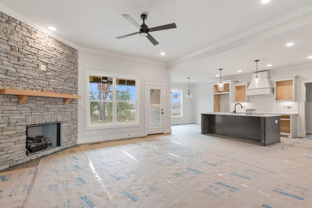 kitchen with crown molding, hanging light fixtures, premium range hood, a stone fireplace, and a kitchen island with sink