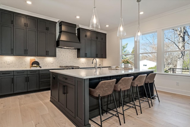 kitchen featuring premium range hood, an island with sink, light wood-type flooring, and decorative light fixtures