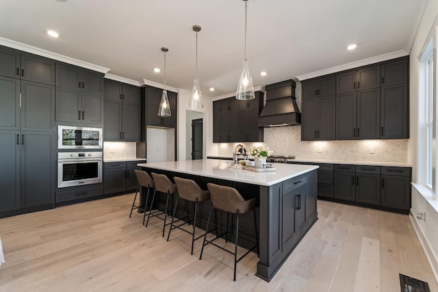 kitchen featuring crown molding, hanging light fixtures, premium range hood, a kitchen island with sink, and stainless steel appliances