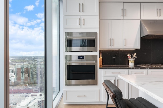 kitchen with wall chimney exhaust hood, white cabinetry, stainless steel appliances, decorative backsplash, and light stone counters