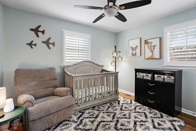 bedroom with a crib, multiple windows, dark wood-type flooring, and ceiling fan