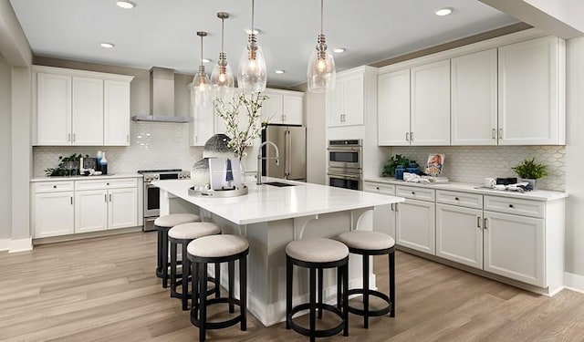 kitchen featuring decorative light fixtures, white cabinetry, an island with sink, stainless steel appliances, and wall chimney exhaust hood