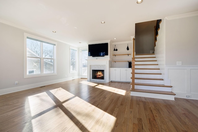 unfurnished living room with a fireplace with flush hearth, wood finished floors, stairs, ornamental molding, and wainscoting