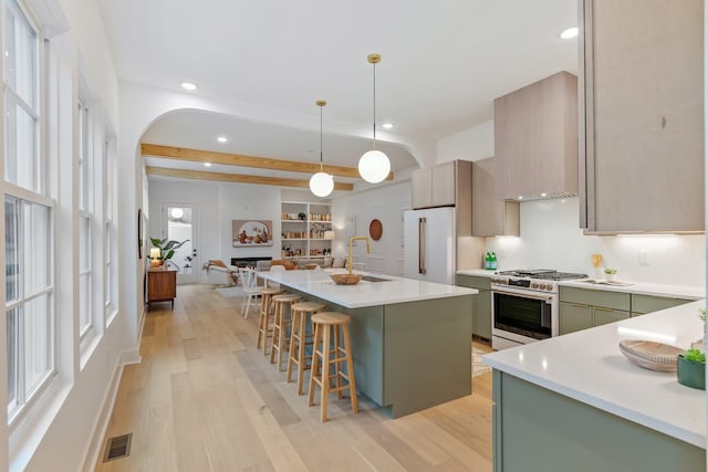 kitchen featuring premium appliances, hanging light fixtures, a kitchen island with sink, green cabinets, and beam ceiling