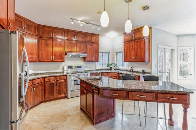 kitchen featuring appliances with stainless steel finishes, hanging light fixtures, a kitchen island with sink, sink, and dark stone counters