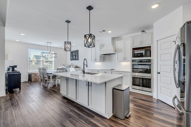 kitchen with hanging light fixtures, premium range hood, white cabinetry, a center island with sink, and black appliances