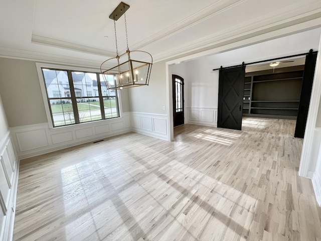 unfurnished dining area featuring a chandelier, light wood-type flooring, a tray ceiling, crown molding, and a barn door