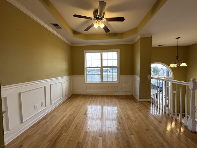 empty room with ceiling fan with notable chandelier, a raised ceiling, ornamental molding, and light hardwood / wood-style floors