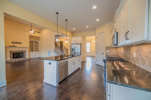 kitchen with appliances with stainless steel finishes, a fireplace, white cabinets, hanging light fixtures, and a kitchen island with sink