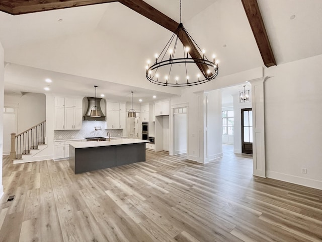 kitchen featuring a kitchen island with sink, white cabinets, decorative light fixtures, wall chimney range hood, and light wood-type flooring