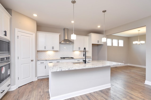 kitchen with white cabinetry, wall chimney range hood, stainless steel appliances, sink, and a kitchen island with sink