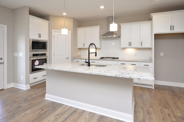 kitchen featuring white cabinets, appliances with stainless steel finishes, wall chimney exhaust hood, an island with sink, and sink