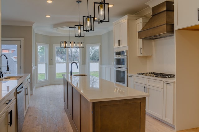 kitchen with appliances with stainless steel finishes, custom range hood, hanging light fixtures, white cabinets, and an island with sink