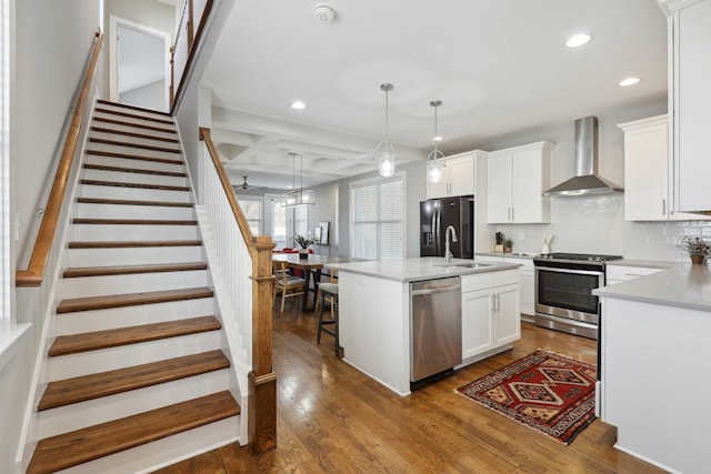 kitchen with wall chimney range hood, white cabinets, decorative light fixtures, stainless steel appliances, and a center island with sink