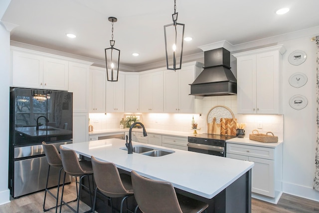 kitchen with white cabinets, custom exhaust hood, sink, stainless steel refrigerator, and a center island with sink