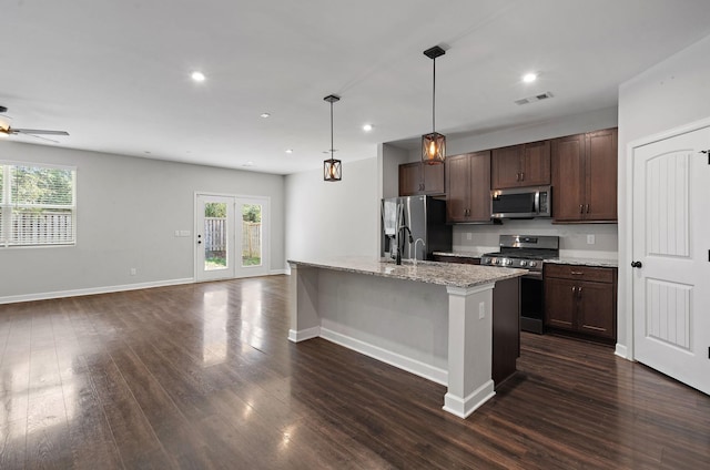 kitchen with light stone countertops, dark brown cabinetry, stainless steel appliances, hanging light fixtures, and a center island with sink